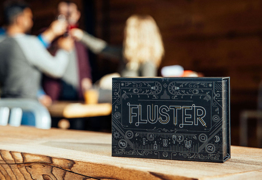 A FLUSTER box on a table with a group of people toasting in the background