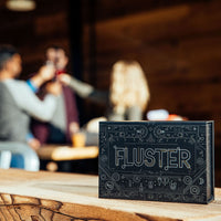 The black Fluster box with intricate design on a bar in the sun in front of a group of people holding up drinks at a brewery.